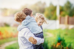 separation or divorce - sleep tips for the family. Photo of an adult outside with grass blurred in the background. The adult si hodling a child in a blue shirt and jeans with short brown hair and another child with longer hair
