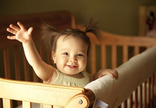 standing in the crib - a smiley baby standing in a light wood sleigh shape crib waving one hand at the camera and the other holding the inside crib rail. Her dark hair is in pigtails.