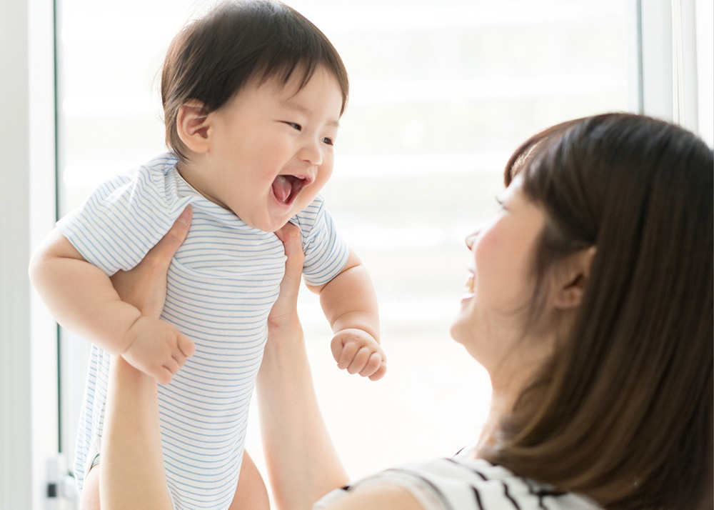regression - a person with shoulder length dark hair wearing a white and navy striped shirt is holding up a baby with short brown hair. The baby is wearing a white and grey striped onesie and is smiling. They are in front of a window that is letting in a lot of light.
