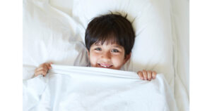 ADHD - a child with dark short hair laying on a white pillow with a white sheet pulled up to his smiling face.