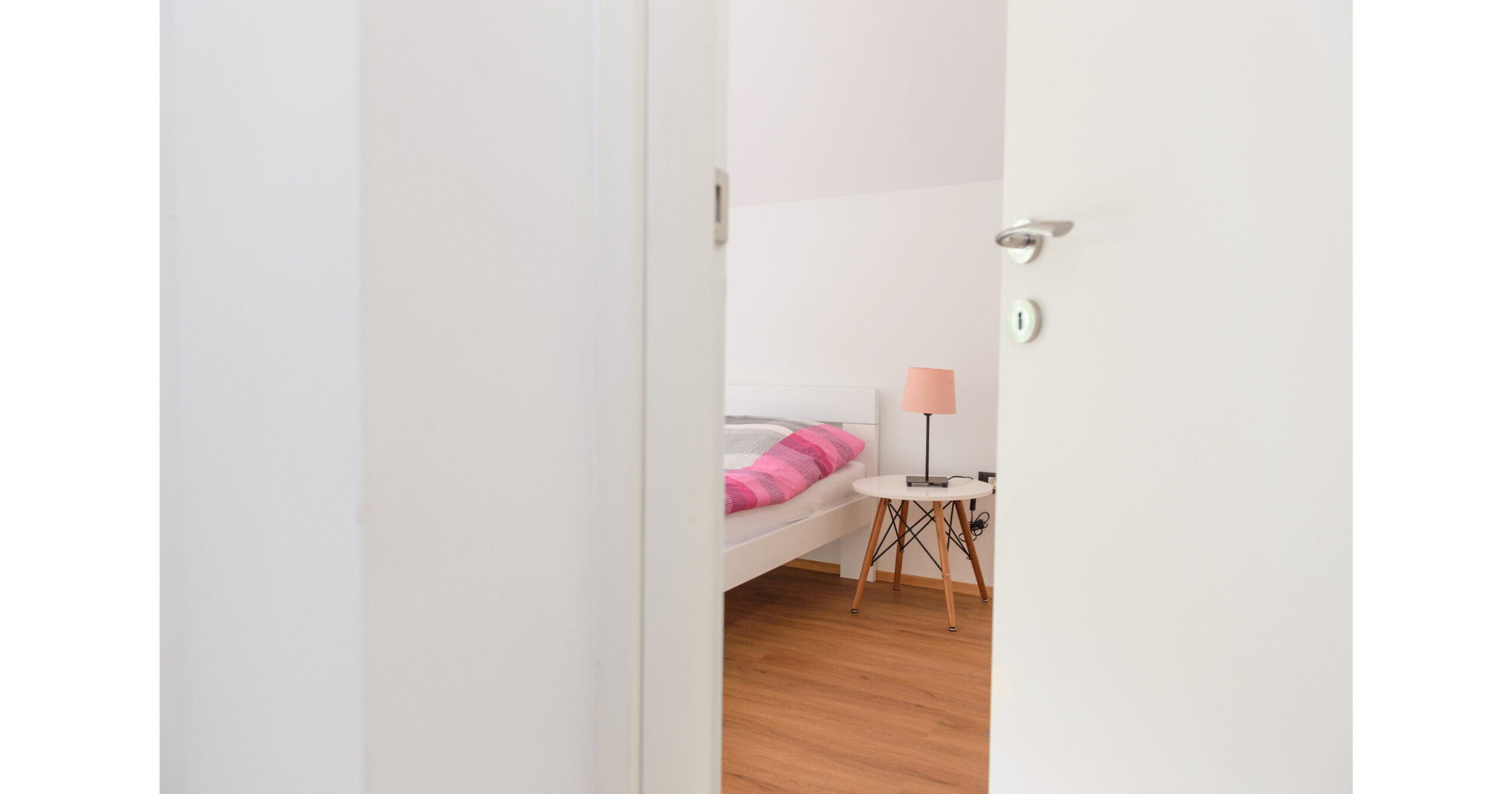 Door to a bedroom partly open with the edge of a bed in view. A light pink lamp on the table