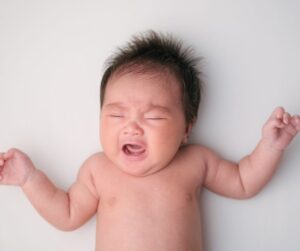 Why Your Baby Cries In Their Sleep And How To Fix It A baby on their back crying with dark hair and eyes closed - arms up beside their head.